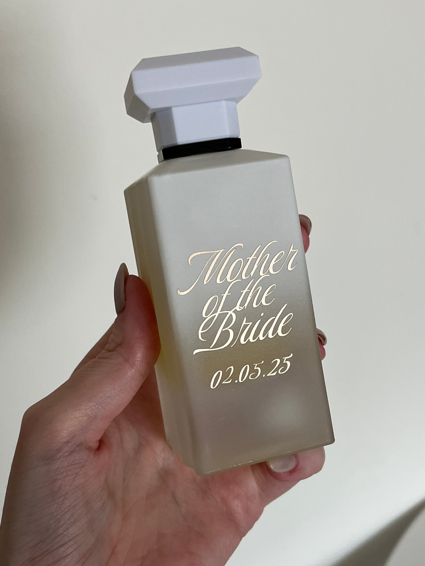 Hand holding a perfume bottle labeled 'Mother of the Bride' with a blurred background