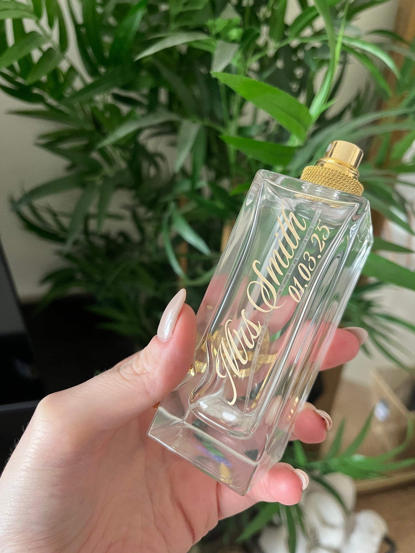 Hand holding a clear perfume bottle with gold cap and custom text against a blurred green plant background