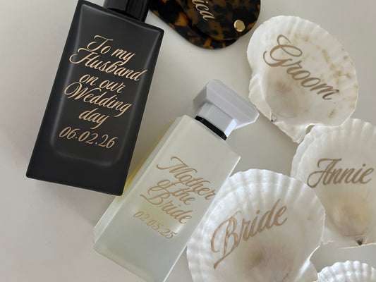 Two perfume bottles with gold text on a white surface surrounded by seashells.