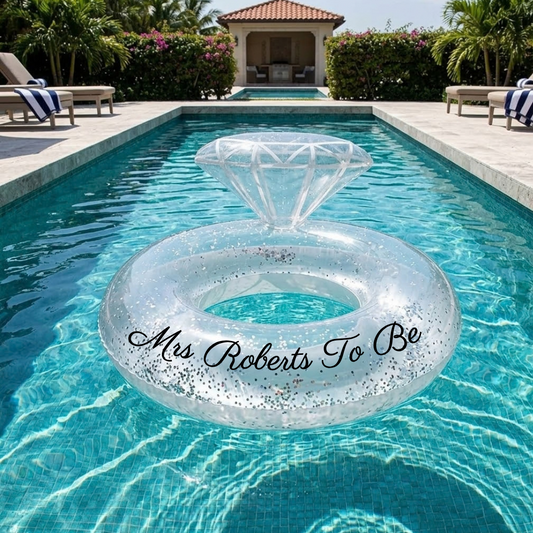 A clear inflatable pool float shaped like a diamond ring with black script text reading 'Mrs Roberts To Be', floating on a swimming pool in a luxurious backyard setting with lounge chairs and a patio umbrella in the background.