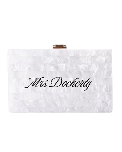 A white textured clutch bridal bag with a small gold metal clasp at the top and the name 'Mrs. Docherty' printed in black cursive across the front, shown on a plain white background.