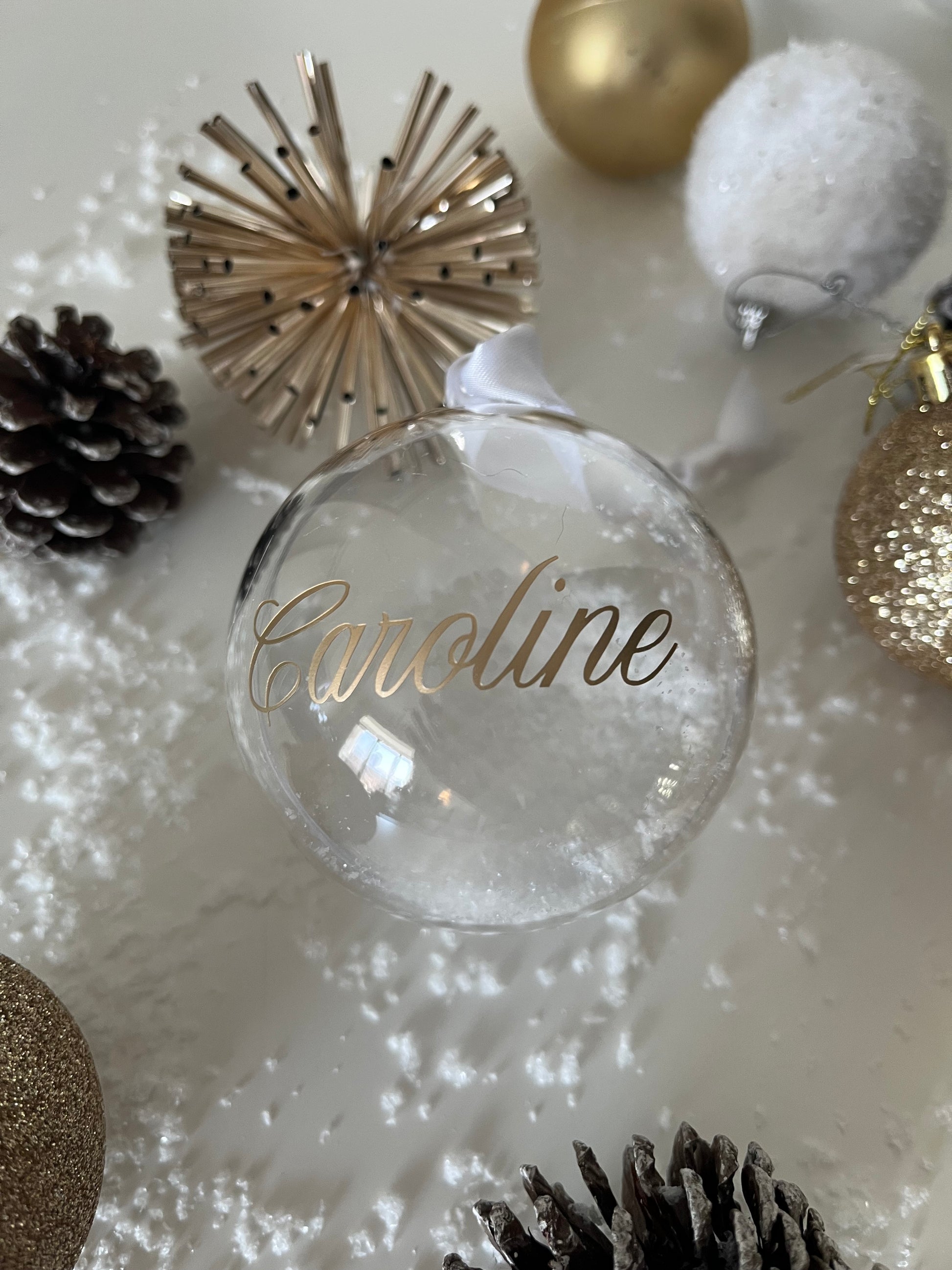 Clear ornament with 'Caroline' engraved, surrounded by Christmas decorations on a white surface.