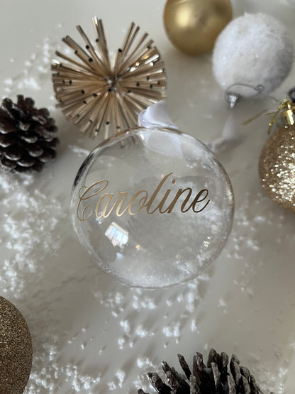 Clear ornament with 'Caroline' engraved, surrounded by Christmas decorations on a white surface.